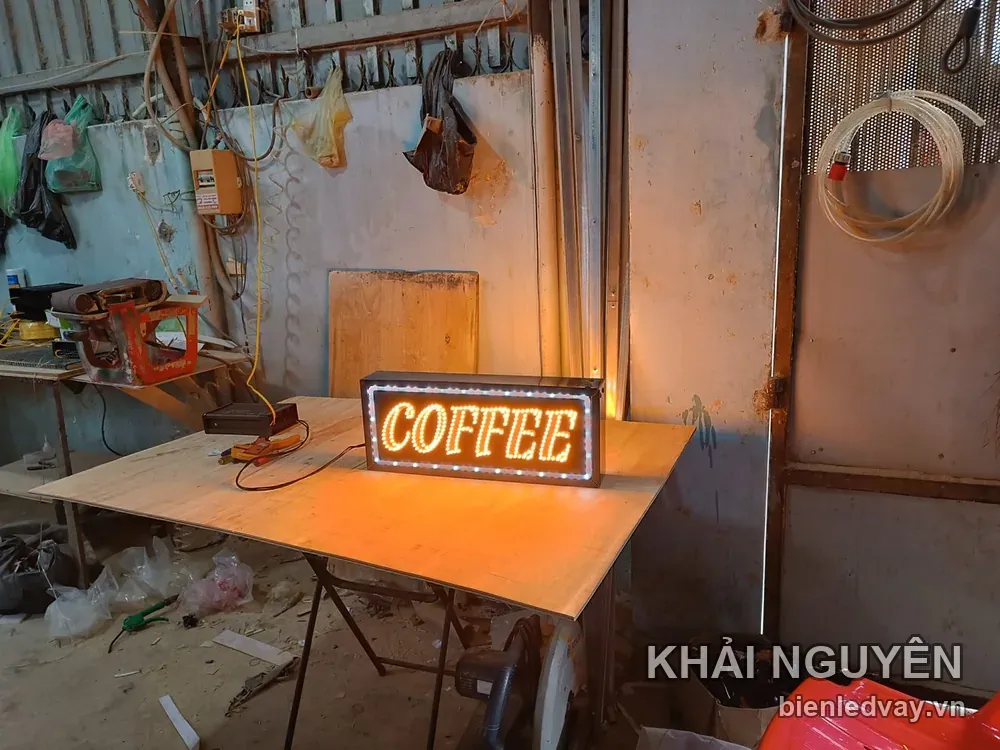 Hình chụp biển vẫy nhỏ có chữ COFFEE sáng vàng cam, viền LED trắng và khung đen, đặt trên bàn gỗ trong xưởng cơ khí.