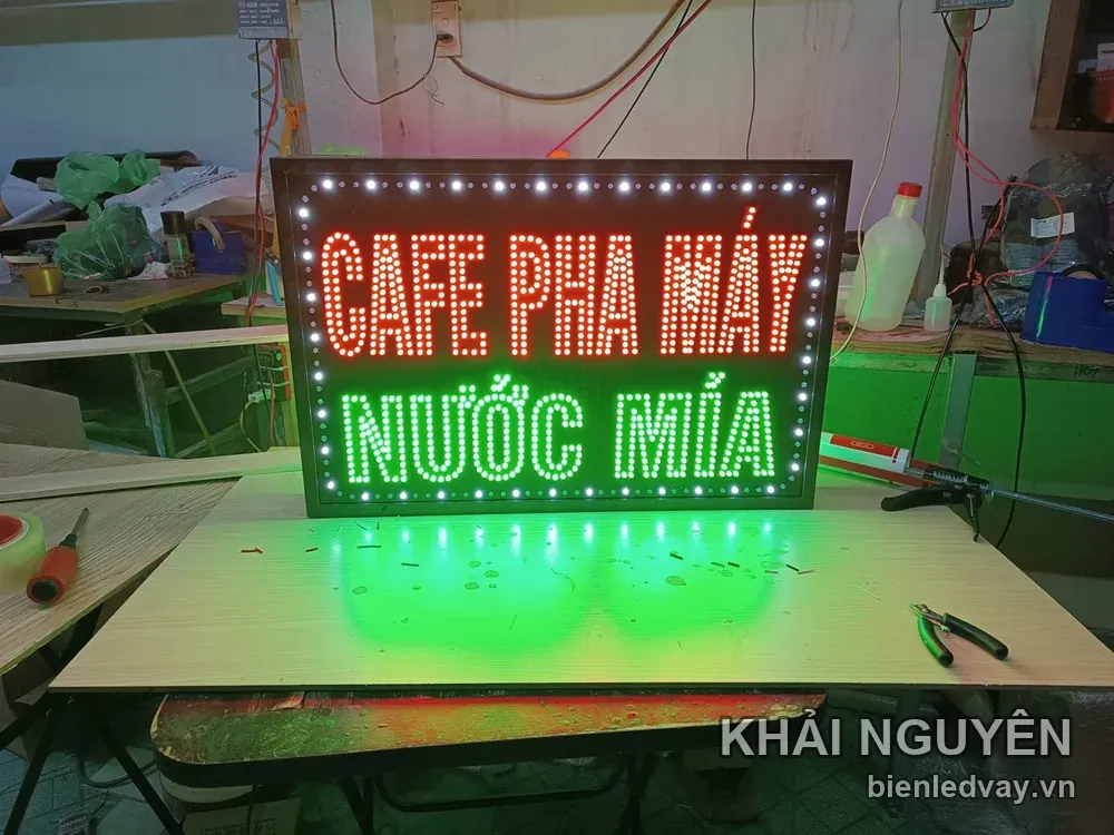 Cận cảnh biển vẫy CAFE PHA MÁY chữ đỏ và NƯỚC MÍA chữ xanh, ánh sáng xanh loang trên mặt bàn cạnh kìm và tua vít