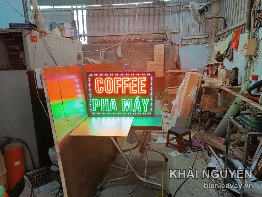 Biển vẫy Coffee Pha Máy có viền bóng tròn trắng, chữ LED cam và xanh lá đang bật sáng thử trong xưởng gỗ.
