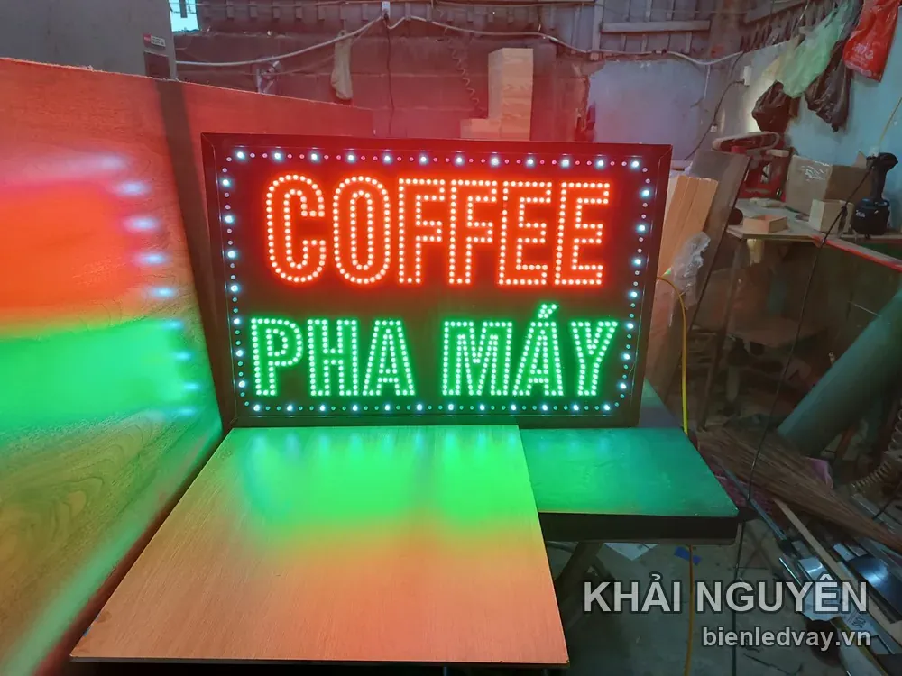 Biển vẫy COFFEE PHA MÁY phát sáng đỏ xanh trong xưởng, có ánh LED hắt lên mặt bàn gỗ và tấm ván bên cạnh.