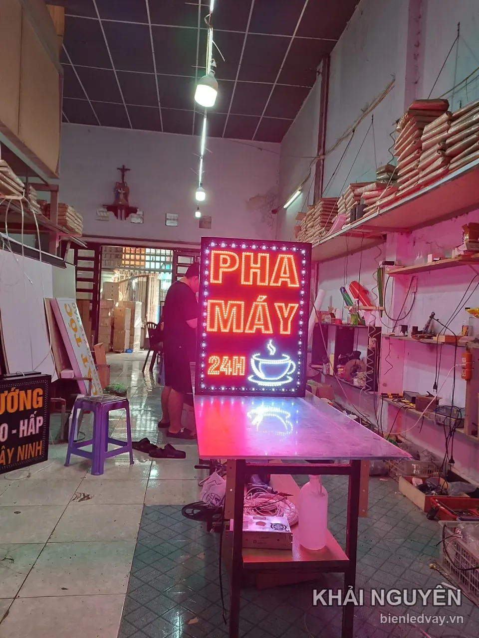Biển vẫy PHA MÁY 24H dáng đứng phát sáng hồng đỏ, chụp trong toàn cảnh xưởng biển có kệ vật tư ở hai bên.