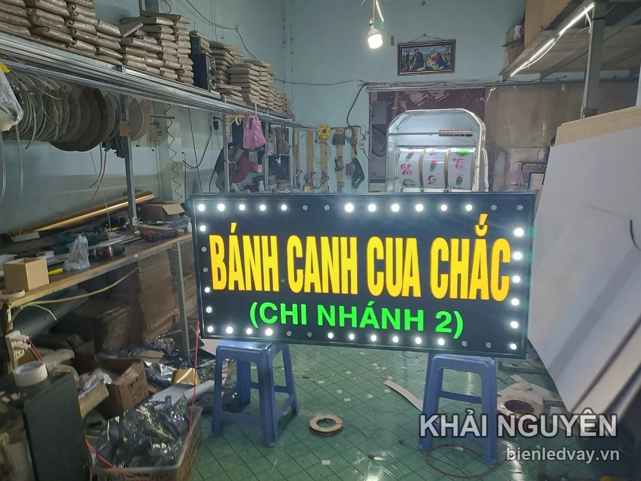 Biển vẫy LED Bánh Canh Cua Chắc chữ vàng, dòng Chi Nhánh 2 xanh lá, viền bóng trắng, đặt trên ghế tại xưởng