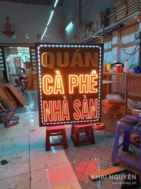 Biển vẫy LED Quán Cà Phê Nhà Sàn chữ cam viền chấm bi trắng, dựng đứng trên hai ghế nhựa trong xưởng
