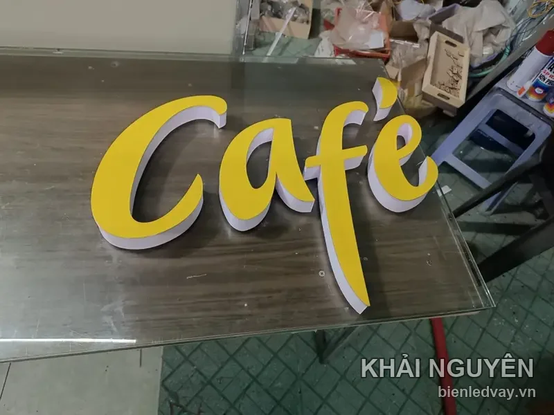 Chữ nổi mica Café kiểu viết tay màu vàng, thành chữ trắng, đặt nằm trên mặt bàn kính trong xưởng chờ lắp