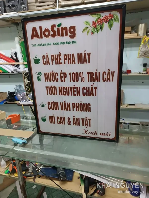 Cận cảnh hộp đèn AloSing chữ đỏ trên nền trắng vân gỗ, logo lá xanh và chùm quả cà phê chín, khung nâu, chân sắt
