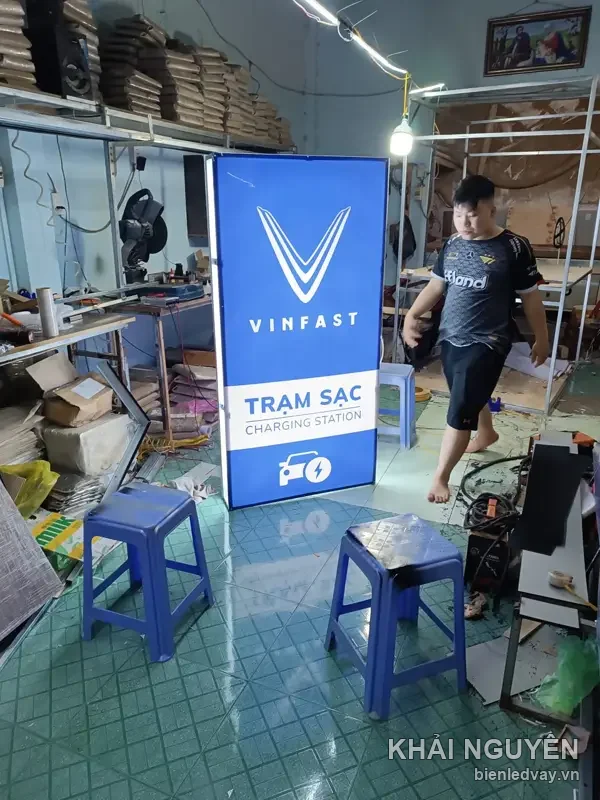 Hộp đèn Trạm Sạc VinFast cao ngang người, mặt xanh in logo V và biểu tượng ô tô sạc, thợ đi ngang trong xưởng