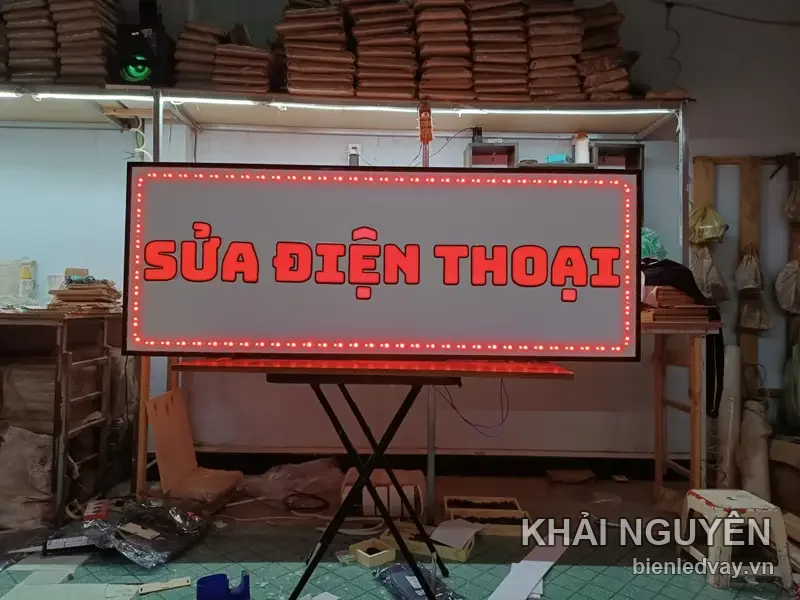 Hộp đèn SỬA ĐIỆN THOẠI chữ đỏ trên nền trắng, viền LED bóng tròn đỏ, đặt trên giá chữ X trong xưởng