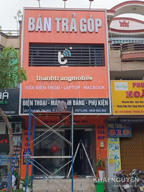 Mặt tiền thanhtrungmobile với bảng alu cam, chữ nổi BÁN TRẢ GÓP trắng, thợ đang lắp trên giàn giáo