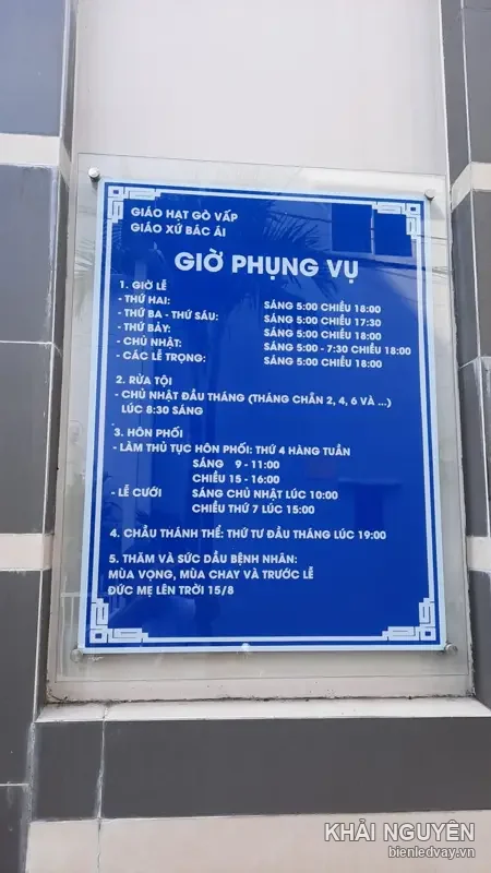 Bảng Giờ Phụng Vụ nền xanh dương chữ trắng của Giáo hạt Gò Vấp - Giáo xứ Bác Ái, gắn trên tường ốp đá