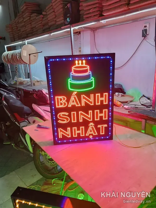 Biển vẫy LED Bánh Sinh Nhật chữ cam viền chấm bi, hình bánh kem ba tầng nhiều màu, dựng trên bàn xưởng