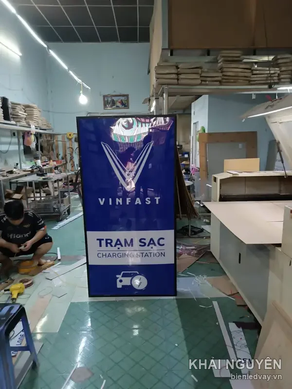 Hộp đèn VinFast nền xanh dương, logo chữ V và dòng TRẠM SẠC - CHARGING STATION nền trắng, dựng trong xưởng