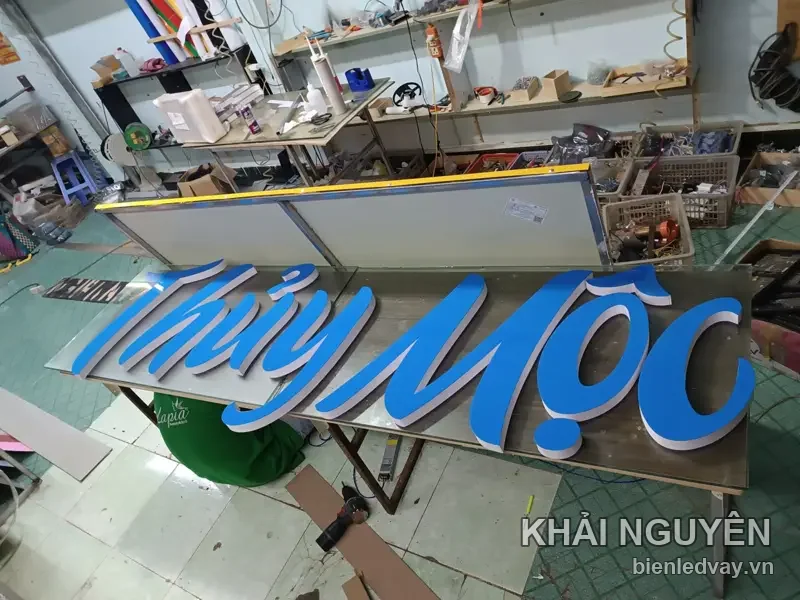 Chữ nổi mica Thủy Mộc kiểu viết tay màu xanh dương gắn trên nền inox bạc, nằm trên bàn lắp ráp tại xưởng