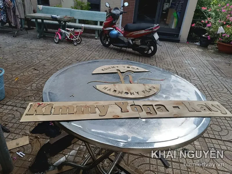Chữ nổi gỗ cắt CNC Kimmy Yoga - Katy cùng logo tròn Harmony Flow, đặt trên mặt bàn inox ngoài sân trước khi lắp
