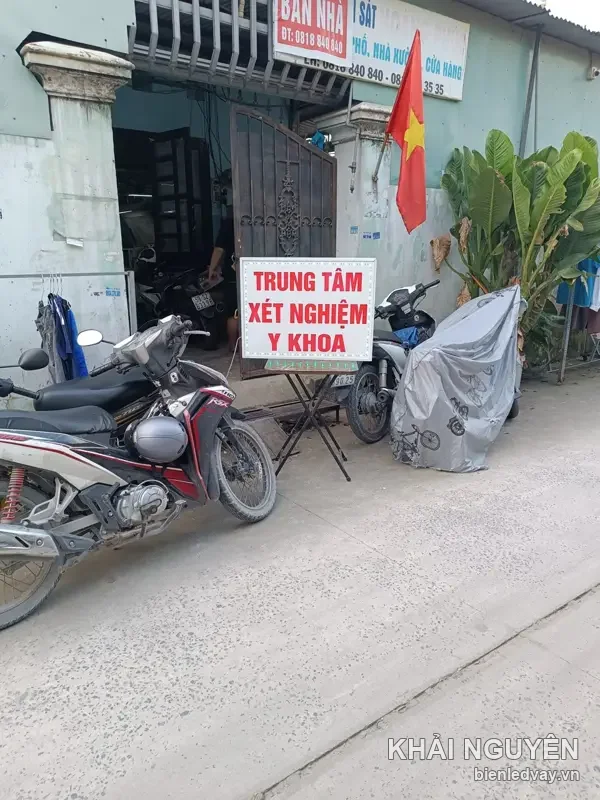 Hộp đèn mica chữ đỏ TRUNG TÂM XÉT NGHIỆM Y KHOA trên giá chữ X, đặt sát lề đường cạnh hàng xe máy