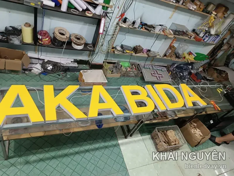 Chữ nổi mica vàng AKA BIDA đặt trên bàn kính trong xưởng, dây điện LED nối sẵn phía sau chờ đấu nguồn