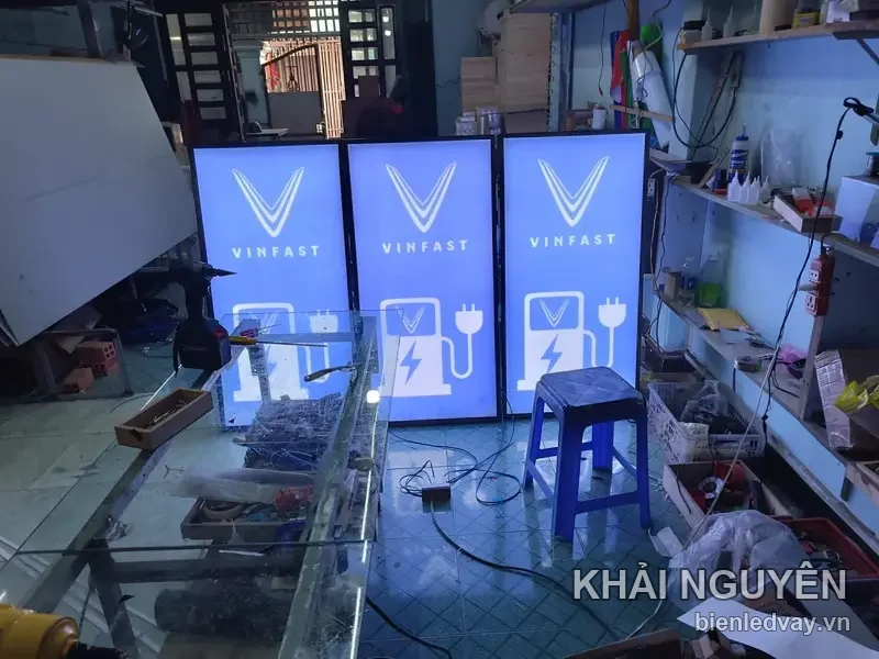 Ba hộp đèn VinFast nền xanh dương, logo chữ V và biểu tượng trụ sạc xe điện màu trắng, đang sáng thử