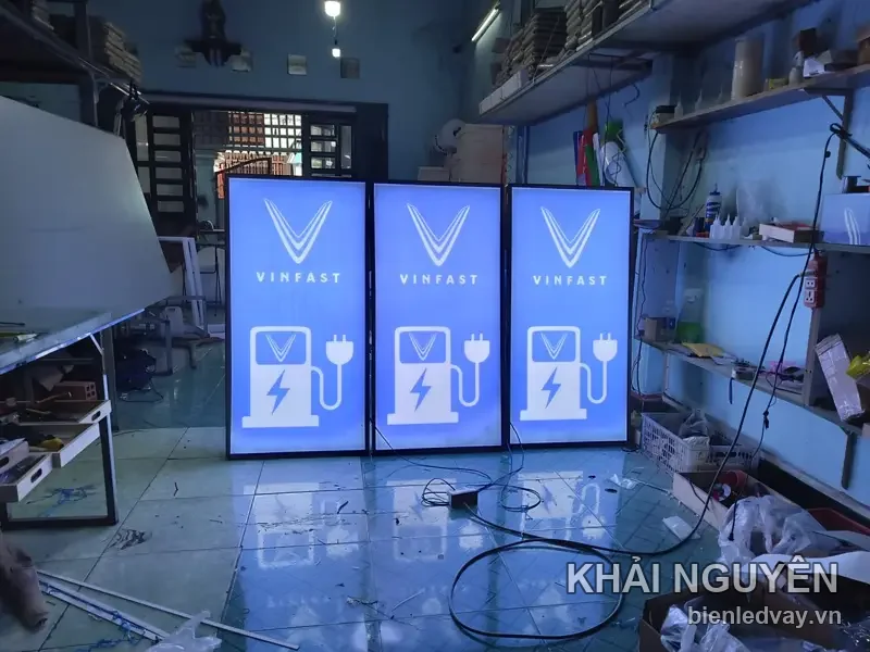 Ba hộp đèn VinFast nền xanh dương, logo chữ V và biểu tượng trụ sạc xe điện màu trắng, đang thử sáng tại xưởng