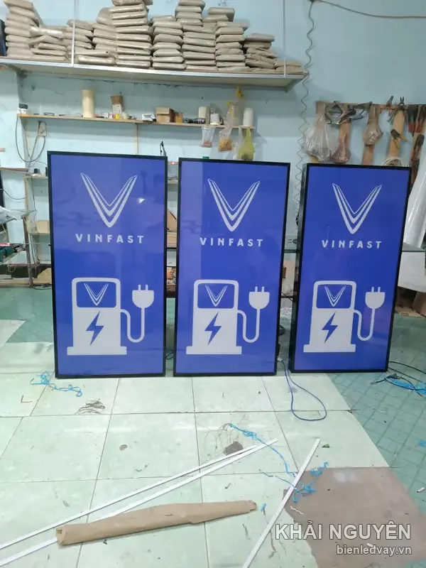 Ba hộp đèn VinFast nền xanh dương, logo chữ V trắng và biểu tượng trụ sạc xe điện, xếp trong xưởng