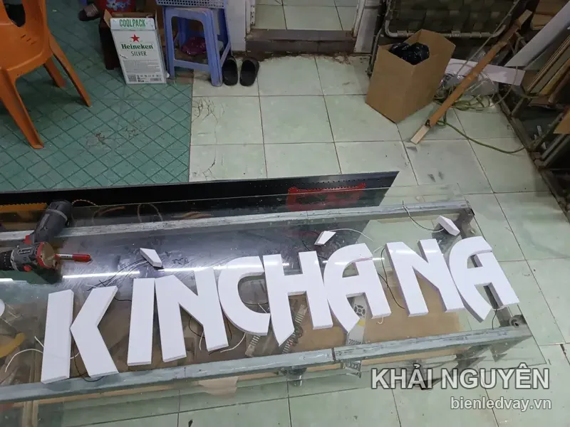 Chữ nổi mica trắng KINCHANA đang bắt lên thanh nhôm, máy khoan và dây LED bày quanh sàn xưởng