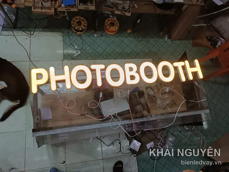 Chữ nổi mica PHOTOBOOTH sáng vàng ấm nằm trên bàn xưởng, dây điện và nguồn LED còn đấu tạm để thử sáng