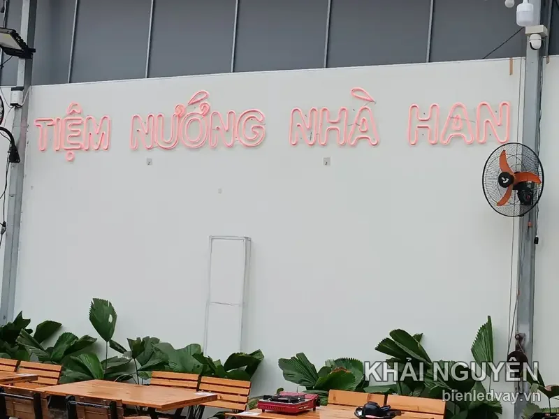 Tiệm Nướng Nhà Han: chữ nổi uốn ống LED neon hồng gắn thẳng lên tường trắng, dưới là bàn ghế gỗ sân quán