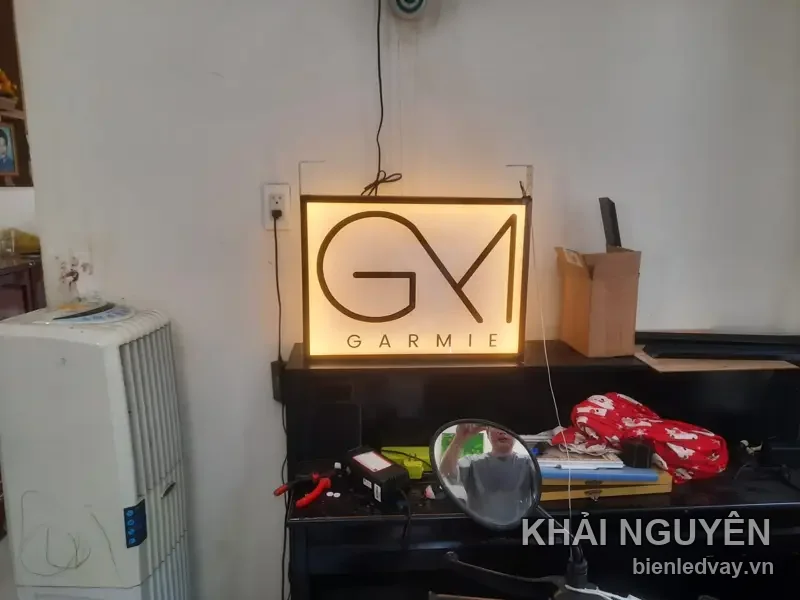 Hộp đèn mica logo GM lồng chữ và tên Garmie, hắt sáng vàng ấm, đặt trên bàn xưởng trước khi giao