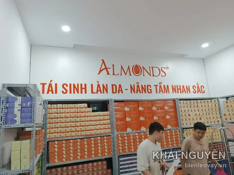 Chữ nổi ALMONDS đỏ gắn tường trắng cùng dòng Tái sinh làn da - Nâng tầm nhan sắc, phía dưới là kệ mỹ phẩm