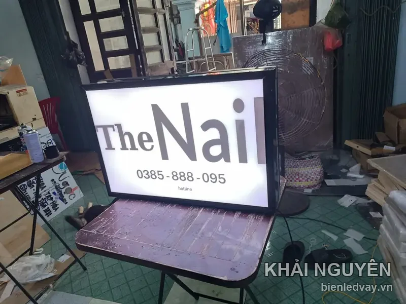 Hộp đèn khung đen The Nail chữ xám trên nền trắng kèm hotline số liên hệ trên biển, đặt trên bàn gấp ở xưởng