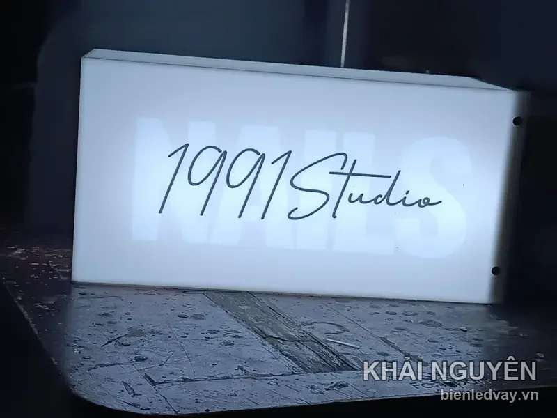 Hộp đèn mica trắng 1991 Studio chữ viết tay màu đen, chữ NAILS chìm phía sau, chụp đêm trên nền bê tông