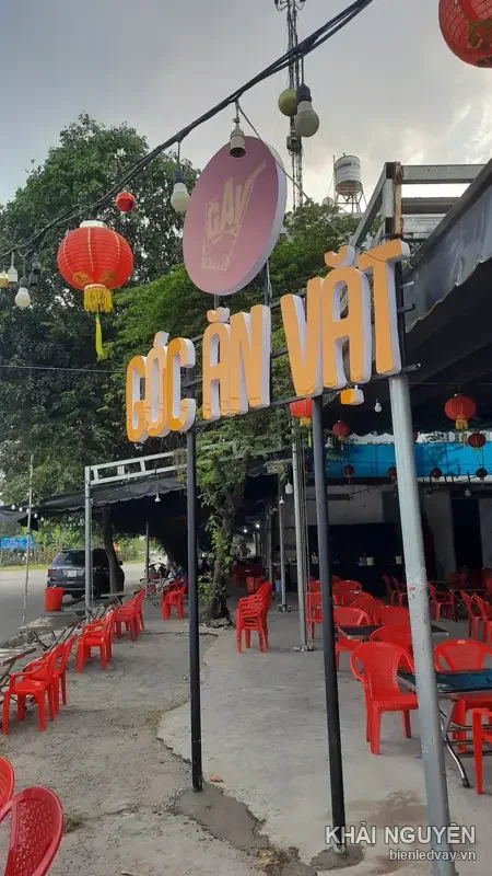 Chữ nổi Góc Ăn Vặt màu vàng nhìn nghiêng, phía sau là bàn ghế nhựa đỏ, đèn lồng đỏ và logo GAV nền hồng