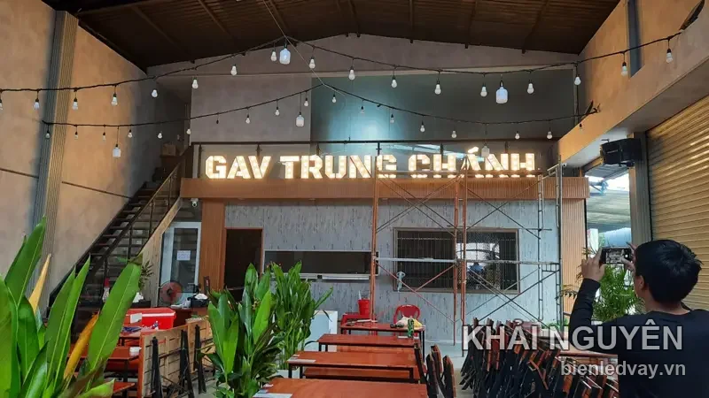 Chữ nổi GAV TRUNG CHÁNH phát sáng vàng ấm trên tường xi măng, dưới dàn đèn dây, quán kê bàn ghế gỗ