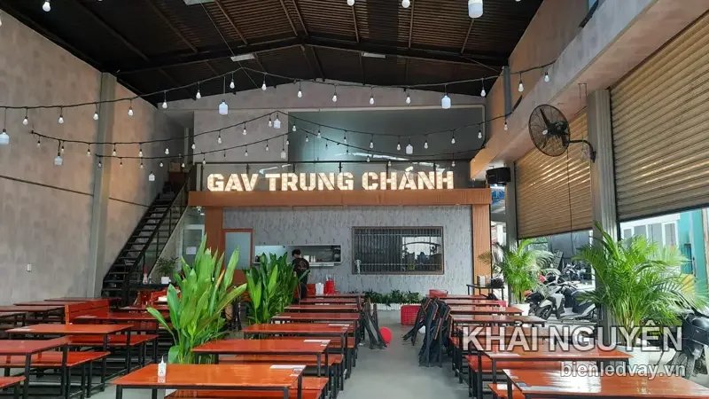 Chữ nổi GAV Trung Chánh màu vàng trên vách sẫm trong quán, phía dưới là bàn ghế gỗ đỏ và dây đèn