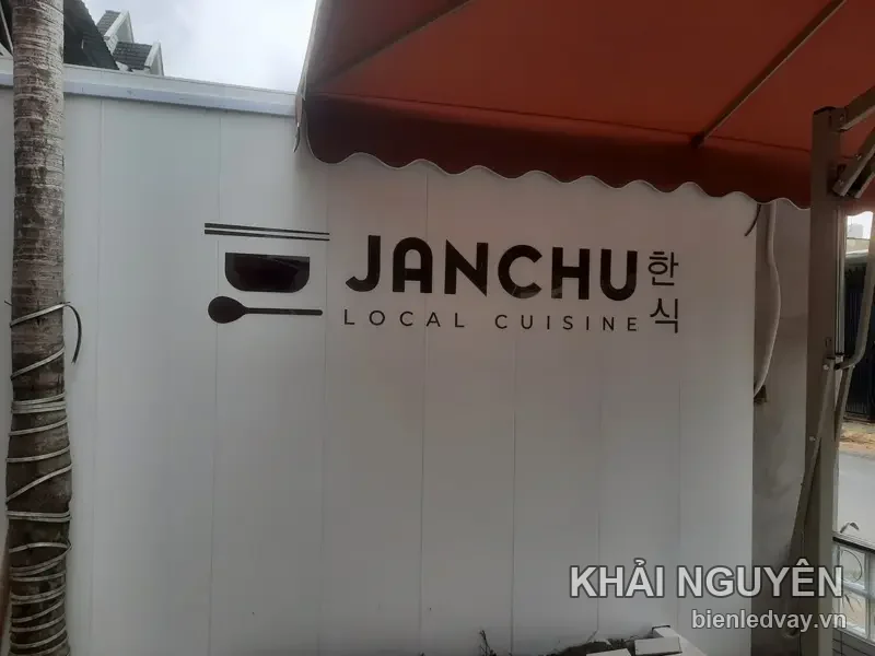 Chữ nổi JANCHU màu nâu đen gắn trên tường tôn trắng, kèm chữ Hàn và dòng LOCAL CUISINE, dưới mái hiên đỏ