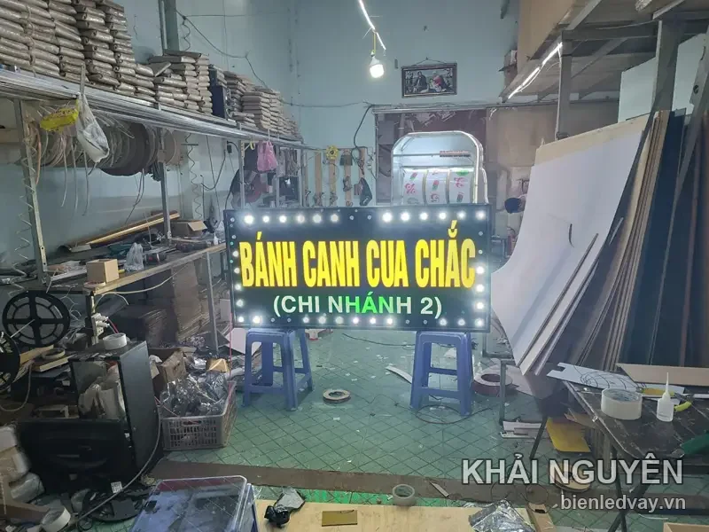Toàn cảnh xưởng Khải Nguyên với biển vẫy BÁNH CANH CUA CHẮC chi nhánh 2 sáng đèn, kê trên hai ghế nhựa
