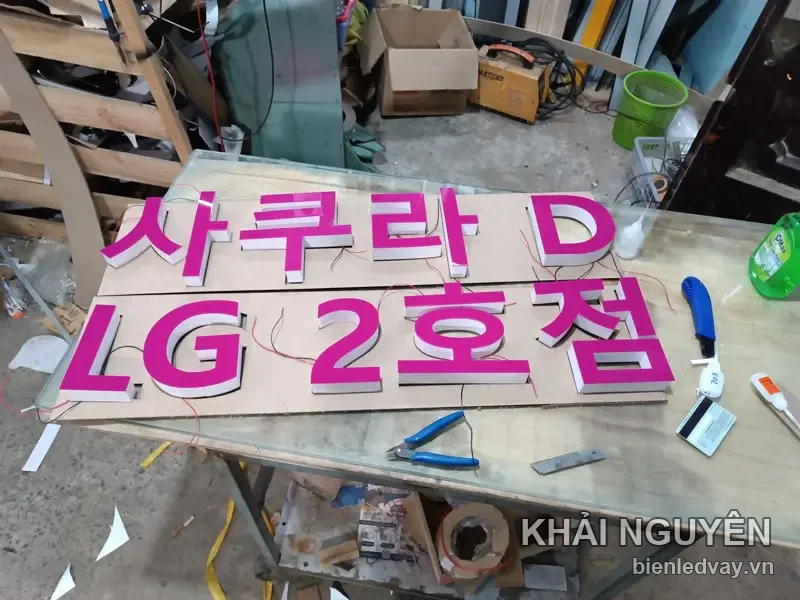 Chữ nổi mica hồng sen tiếng Hàn Sakura LG 2호점, chân chữ trắng, dây điện đỏ chờ đấu, đặt trên bàn gỗ xưởng