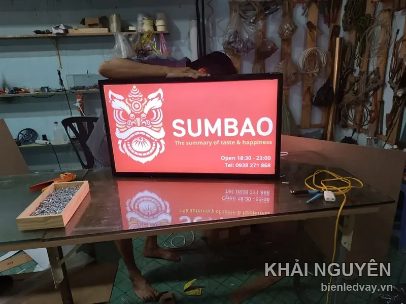 Hộp đèn SUMBAO nền đỏ với hình mặt lân trắng, kèm dòng giờ mở cửa và số điện thoại, đặt trên bàn xưởng