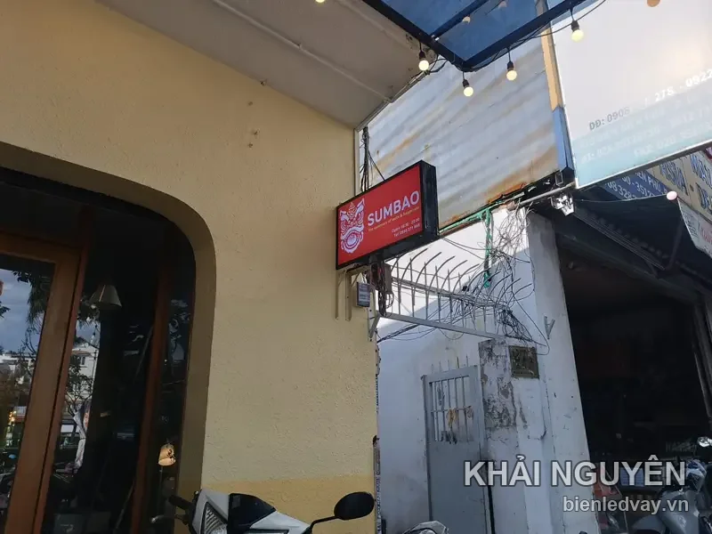 Hộp đèn SUMBAO nền đỏ, logo đầu lân trắng, gắn vuông góc tường vàng trước quán, dây đèn bóng tròn phía trên