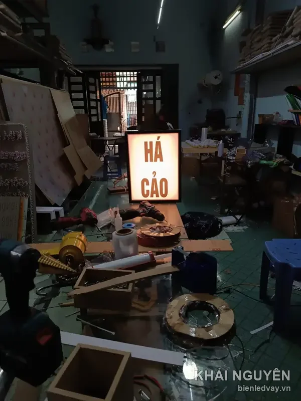 Hộp đèn chữ Há Cảo màu đỏ trên nền vàng sáng, khung viền đen, dựng giữa xưởng ngổn ngang vật tư