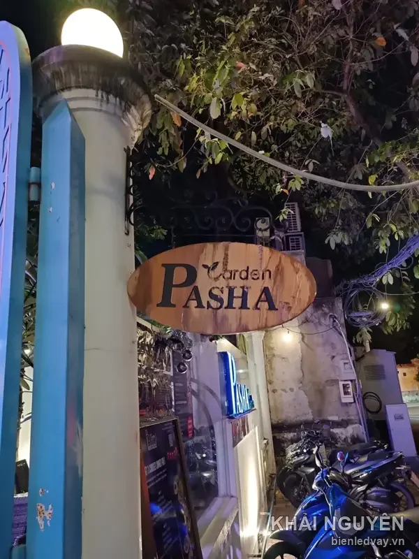 Biển gỗ oval Garden PASHA chữ đen treo ở cổng nhà hàng buổi tối, phía trong có hộp đèn nhỏ màu xanh