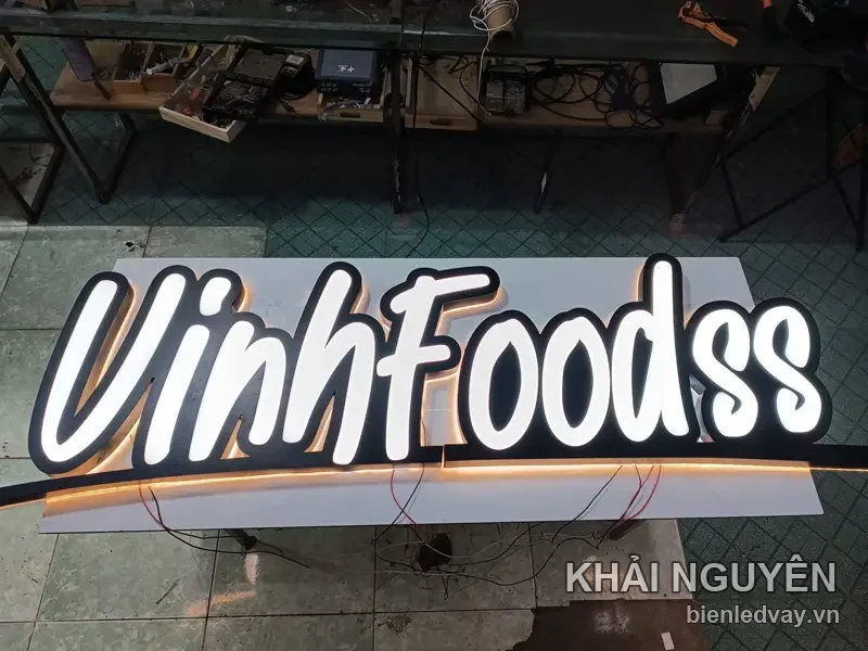 Chữ nổi mica VinhFoodss kiểu chữ viết tay, mặt trắng viền đen, hắt LED vàng, đặt trên nền xưởng