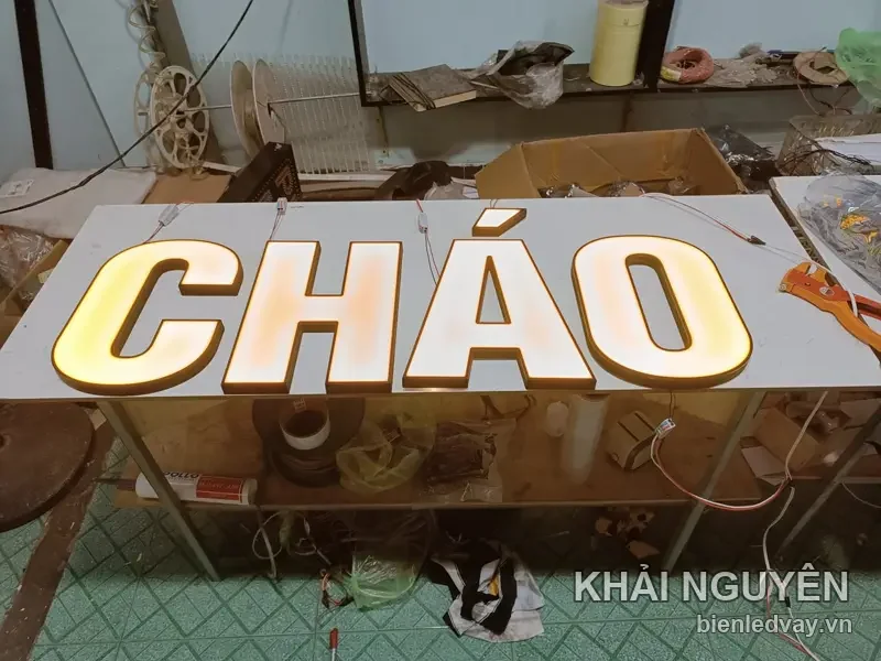 Chữ nổi mica CHÁO cỡ lớn, mặt trắng thành vàng đồng, hắt sáng vàng ấm, đặt trên bàn xưởng