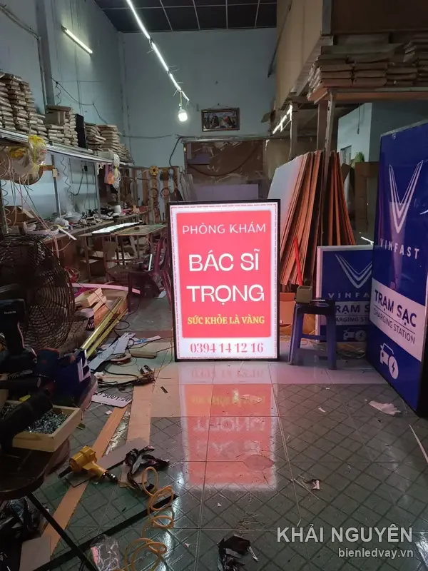 Hộp đèn Bác Sĩ Trọng mặt mica đỏ rực chữ trắng, dựng giữa lối đi xưởng, ánh sáng hắt loang xuống nền gạch
