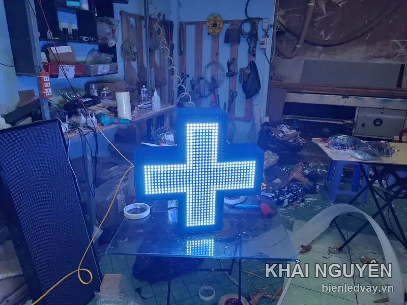 Đèn chữ thập LED nhà thuốc dạng hộp nổi, mặt kín bóng LED trắng ấm, thân hộp xanh dương, đặt trên bàn kính xưởng