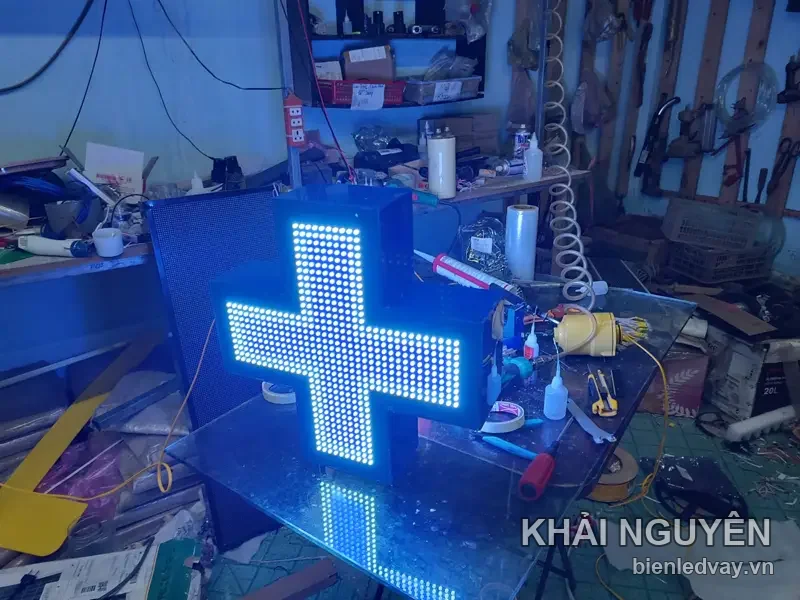 Hộp đèn chữ thập LED nhà thuốc sáng trắng ấm, thân hộp xanh dương, phản chiếu trên mặt bàn kính giữa xưởng