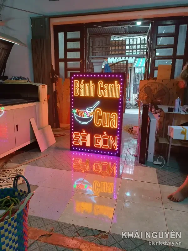 Biển vẫy LED chữ Bánh Canh Cua Sài Gòn cam vàng kèm hình tô bún, viền bóng hồng, dựng gần cửa xưởng
