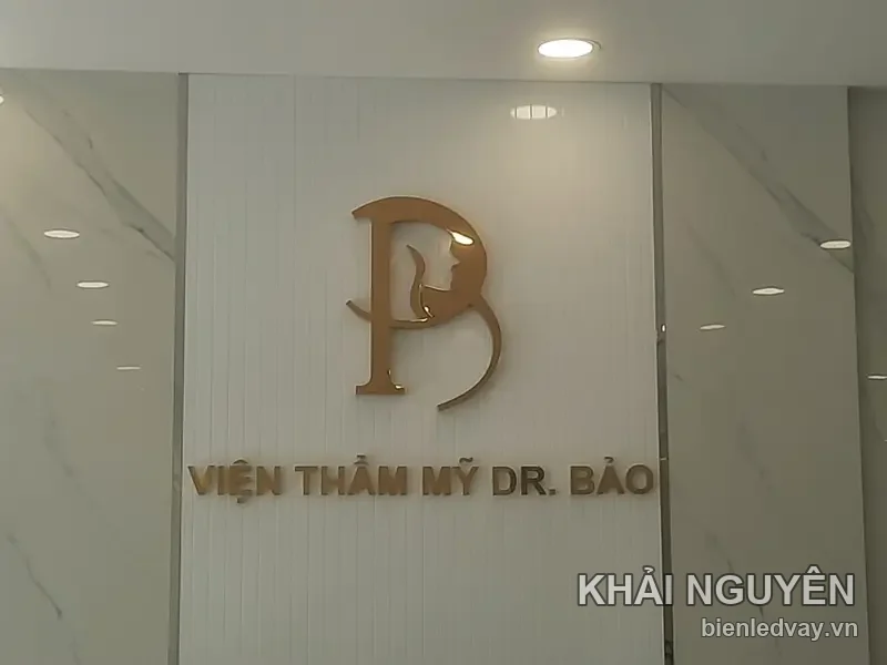 Chữ nổi inox vàng đồng VIỆN THẨM MỸ DR. BẢO cùng logo chữ B lồng gương mặt, gắn trên tường ốp trắng