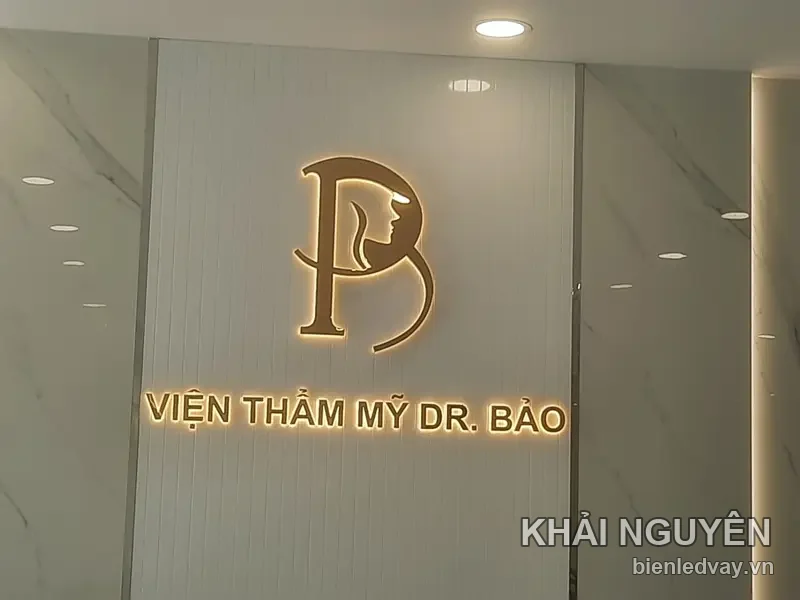 Logo chữ nổi inox vàng Viện Thẩm Mỹ Dr. Bảo với biểu tượng gương mặt cô gái, đèn hắt vàng trên vách đá trắng