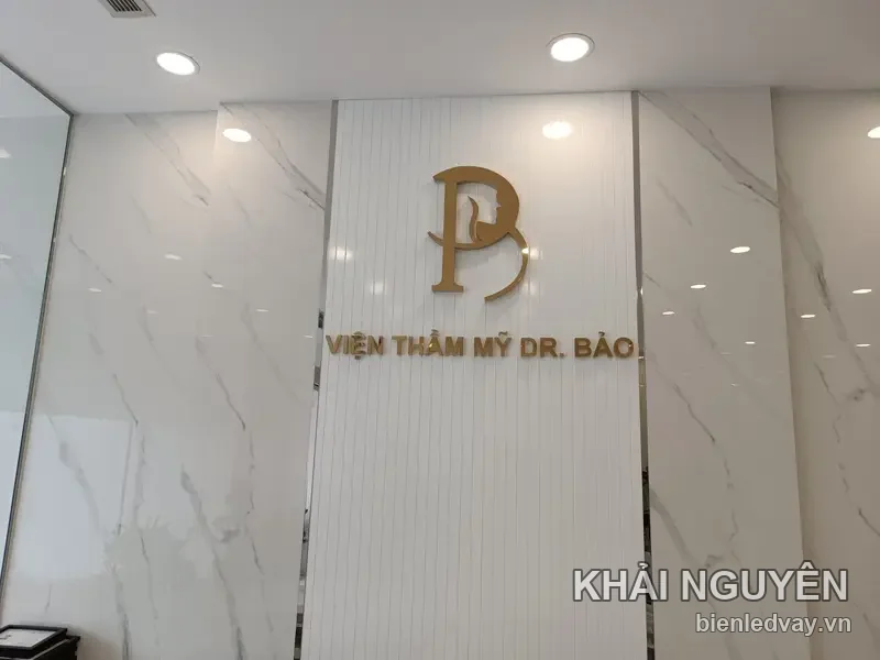 Chữ nổi inox mạ vàng Viện Thẩm Mỹ Dr. Bảo cùng logo chữ B, gắn trên tường vân đá trắng ở sảnh
