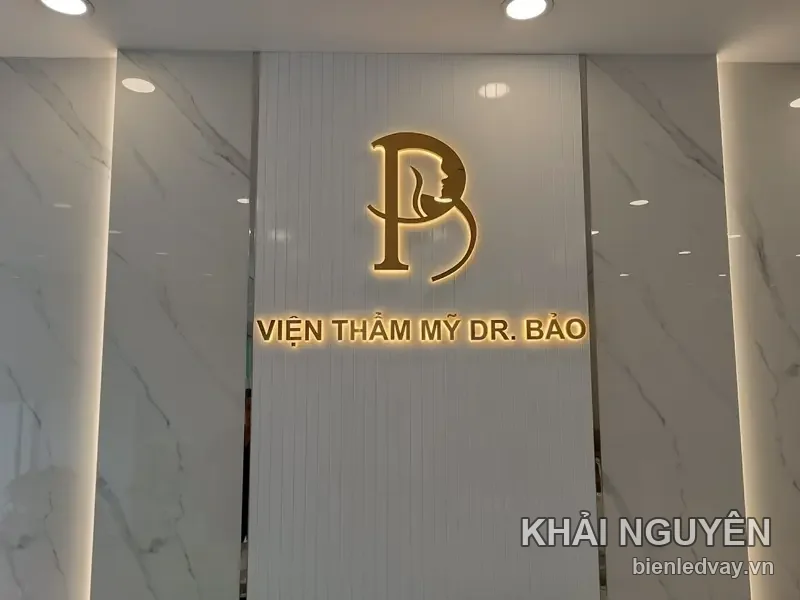 Logo chữ B lồng gương mặt và dòng Viện Thẩm Mỹ Dr. Bảo bằng chữ nổi inox vàng trên tường vân đá trắng