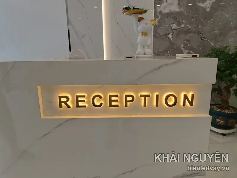 Chữ nổi inox vàng RECEPTION gắn hộc âm trên quầy lễ tân ốp đá trắng, đèn hắt vàng phía sau